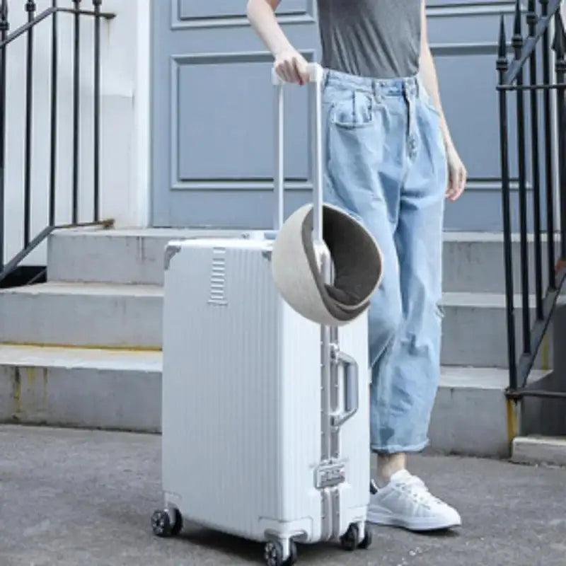 Person holding a white suitcase with a travel neck pillow carrier on top, standing on a sidewalk.